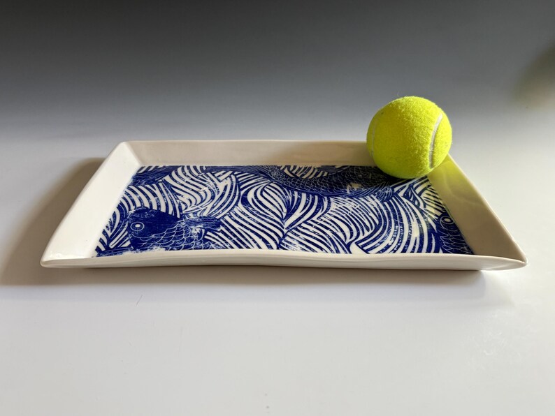 Pu&ograve; includere: Un vassoio rettangolare in ceramica con base bianca e una stampa grafica blu raffigurante pesci stilizzati e motivi a onde. Una pallina da tennis giallo brillante poggia sul lato destro del vassoio, fornendo una scala visiva delle dimensioni.