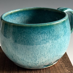 May include: A handmade ceramic mug with a rounded body and a teal glaze. The mug has a handle and sits on a wooden surface. The glaze transitions from a darker teal at the rim to a lighter, textured turquoise towards the base.