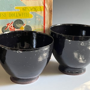 May include: Two black ceramic bowls with a speckled glaze. The bowls have a rounded shape and a small foot.