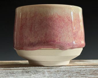 May include: A handmade ceramic cup with a unique design. The upper portion features a blend of pink and cream hues, while the base is a solid cream color. The cup's shape is slightly tapered, adding to its modern aesthetic.