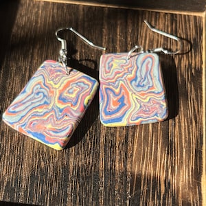 May include: A pair of square earrings with a marbled design in shades of blue, yellow, red, and white. The earrings have silver hooks.