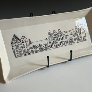 May include: A white ceramic serving tray with a black and red illustration of a row of houses. The tray has a slightly curved shape and is displayed on a black metal stand.