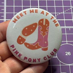 May include: A white button with the text "MEET ME AT THE PINK PONY CLUB" in orange and pink lettering. The button features a pink and orange cowboy boot with stars on the boot.