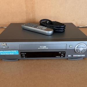 May include: A grey JVC VCR player with a T-V LINK B.E.S.T. PICTURE SYSTEM label, a blue sticker indicating Satellite Controller Ready, a black power cord, and a grey remote control. The front panel features a digital display and playback buttons.