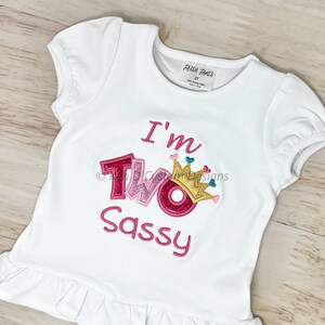 May include: White t-shirt with pink and gold glitter lettering that says "I'm Two Sassy" with a pink crown on top of the "O".