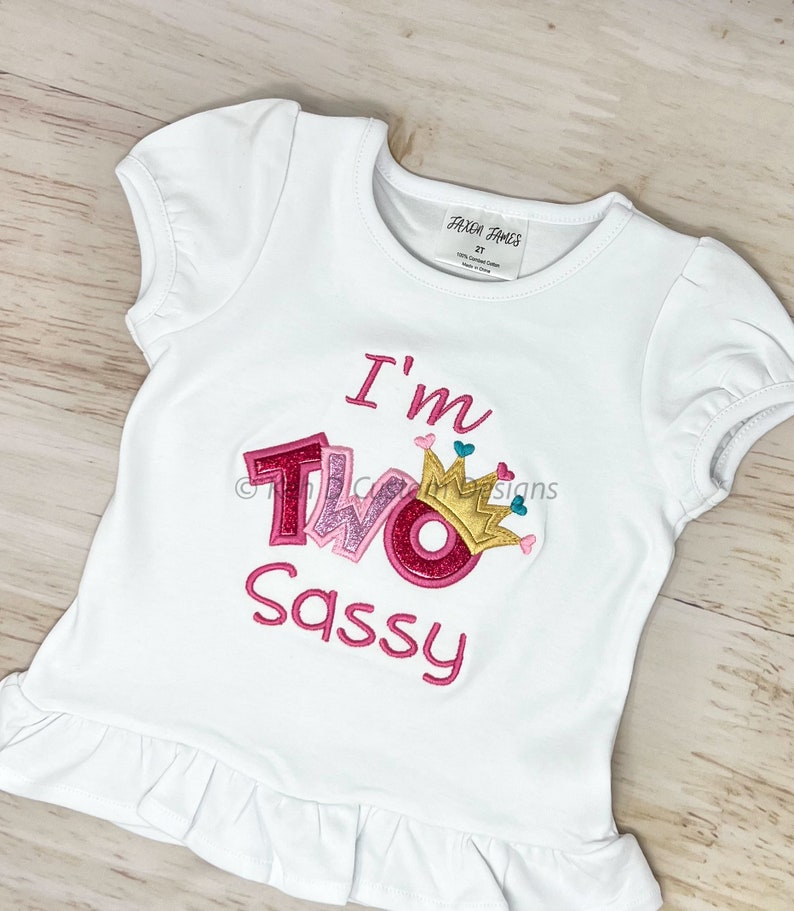 May include: White t-shirt with pink and gold glitter lettering that says "I'm Two Sassy" with a pink crown on top of the "O".