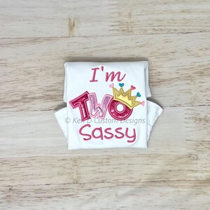 May include: White onesie with pink and gold glitter lettering that says "I'm Two Sassy" with a gold crown and pink hearts above the word "Two".
