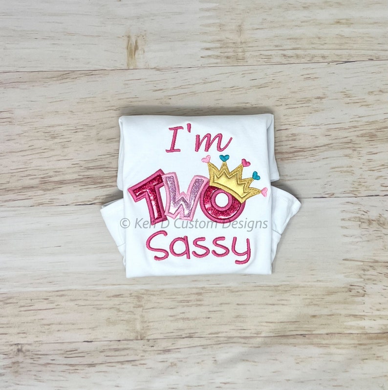 May include: White onesie with pink and gold glitter lettering that says "I'm Two Sassy" with a gold crown and pink hearts above the word "Two".