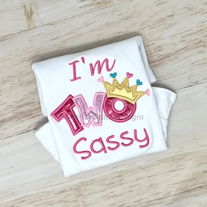 May include: White onesie with pink embroidered text that reads "I'm Two Sassy" with a pink and gold crown above the word "Two".