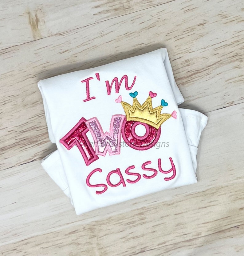 May include: White onesie with pink embroidered text that reads "I'm Two Sassy" with a pink and gold crown above the word "Two".