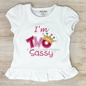 May include: White toddler t-shirt with pink, gold, and teal embroidery. The shirt says "I'm Two Sassy" with a crown above the word "Two".