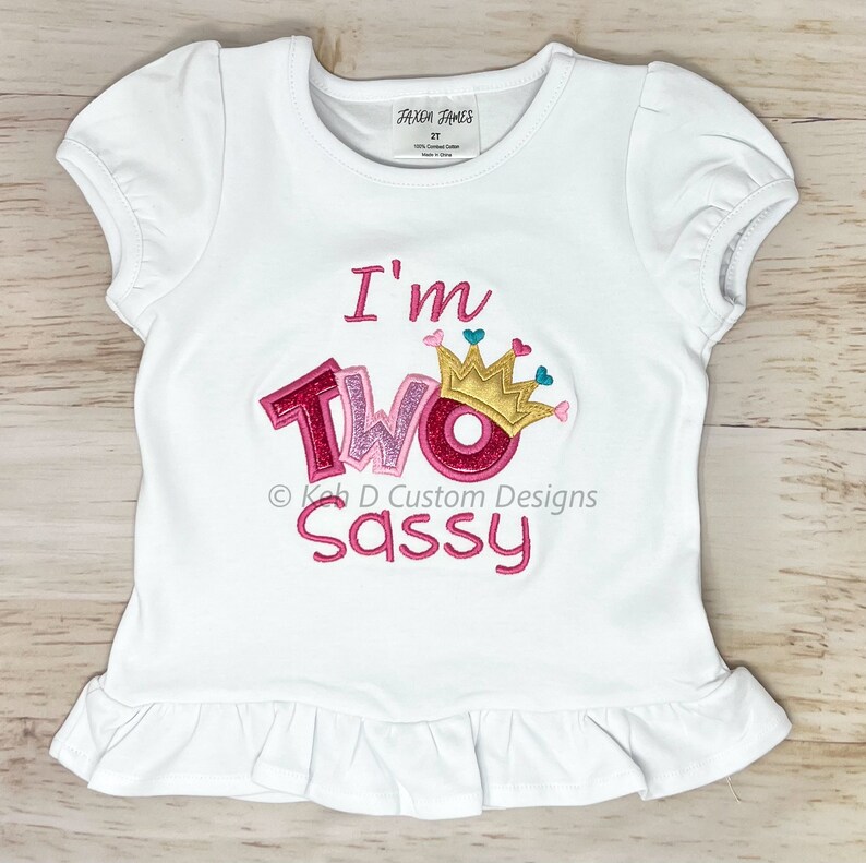 May include: White toddler t-shirt with pink, gold, and teal embroidery. The shirt says "I'm Two Sassy" with a crown above the word "Two".