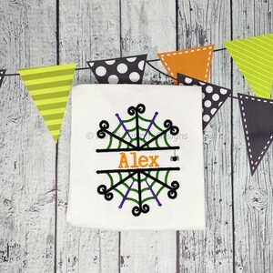 May include: A white t-shirt featuring a black spiderweb design with purple and green accents. The name "Alex" is embroidered in orange text. The shirt is surrounded by colourful pennant flags.