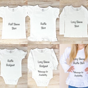 May include: Five different styles of white baby shirts and onesies. The shirts include puff sleeve, ruffle sleeve, and long sleeve options. The onesies include a ruffle and long sleeve option. The text on the shirts reads "Puff Sleeve Shirt", "Ruffle Shirt", "Long Sleeve Shirt", "Ruffle Bodysuit", "Long Sleeve Bodysuit", and "Long Sleeve Ruffle Shirt".