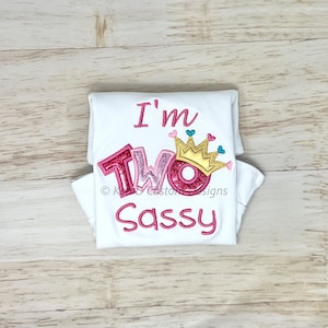 May include: White folded t-shirt with pink and gold embroidery that says "I'm Two Sassy" with a crown and hearts.