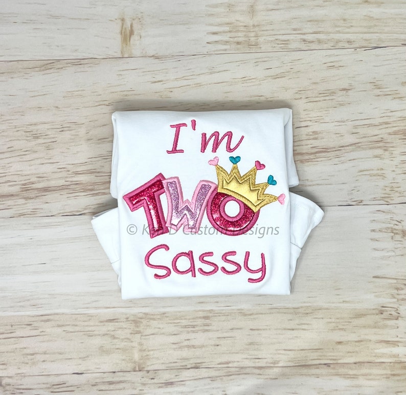 May include: White folded t-shirt with pink and gold embroidery that says "I'm Two Sassy" with a crown and hearts.