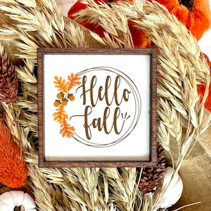 May include: A small square wooden sign with the words "Hello fall" in a circular design. The sign is surrounded by a brown wooden frame and decorated with fall foliage. The background includes dried wheat, orange accents, and white pumpkins.