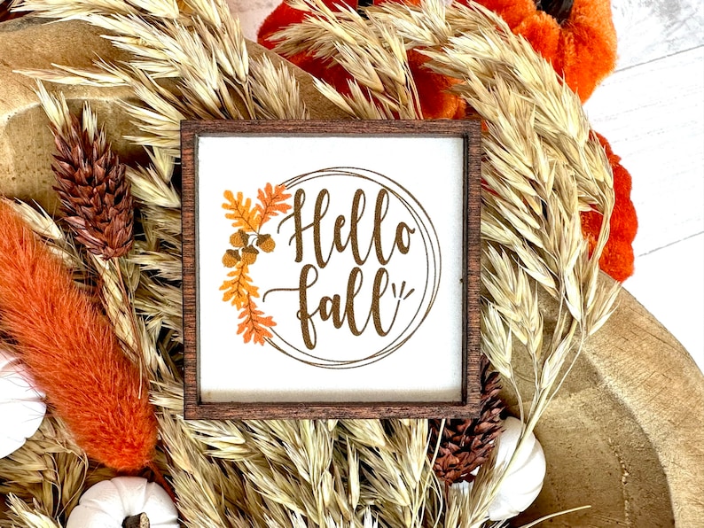 May include: A small square wooden sign with the words "Hello fall" in a circular design. The sign is surrounded by a brown wooden frame and decorated with fall foliage. The background includes dried wheat, orange accents, and white pumpkins.