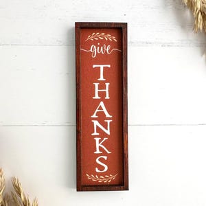 May include: A rectangular wooden sign with a dark brown frame and a burnt orange background. The sign features the white text "give" in a script font above the word "THANKS" in a bold serif font, accented by white leaf illustrations.