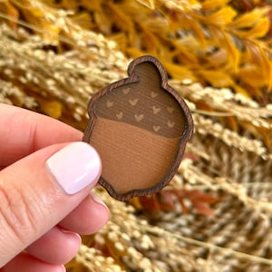 May include: A wooden acorn-shaped pin with a dark brown outline. The top portion is a darker brown with small heart-shaped cutouts, while the bottom is a lighter brown. The pin is held in front of a blurred background of dried plants.