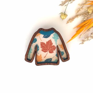 May include: A wooden sweater ornament with a fall leaf pattern. The sweater is brown with a teal, orange, and brown leaf design.