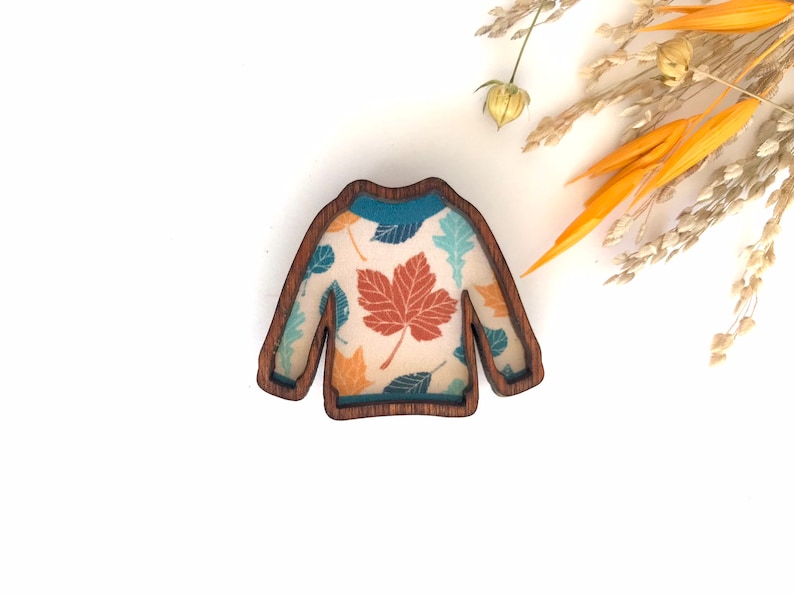 May include: A wooden sweater ornament with a fall leaf pattern. The sweater is brown with a teal, orange, and brown leaf design.