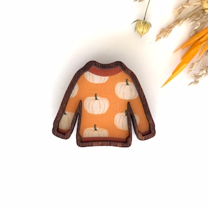 May include: A wooden cutout of a sweater with a pumpkin pattern. The sweater is orange with white pumpkins and has a brown wood frame.