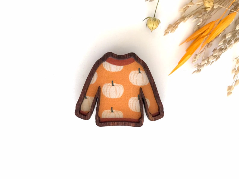 May include: A wooden cutout of a sweater with a pumpkin pattern. The sweater is orange with white pumpkins and has a brown wood frame.