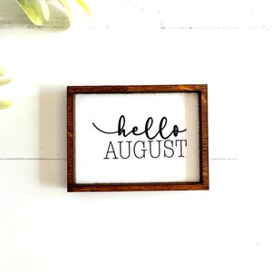 May include: A small wooden sign with a white background and a brown frame. The sign says "hello august" in black cursive lettering.