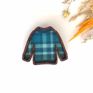 May include: A wooden pin shaped like a sweater with a blue and green plaid pattern.