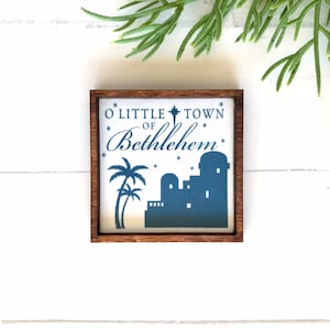 May include: A small wooden framed sign with a white background and blue text and silhouette. The text reads "O Little Town of Bethlehem" with a star and palm trees.