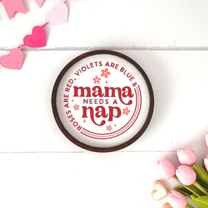 May include: A round wooden sign with the text "Roses are red, violets are blue & mama needs a nap" in red and white. The sign is surrounded by a brown border. Pink heart-shaped garland and pink tulips are in the background.