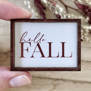 May include: A small wooden sign with a white background and a brown frame. The sign says "hello fall" in copper-colored lettering.