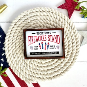 May include: A wooden sign with a red, white, and blue design that says "Uncle Sam's Fireworks Stand". The sign is on a white rope background. The sign also says "Open Daily" and "Made in America".