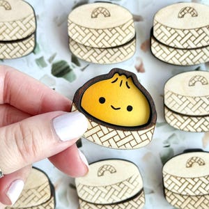 May include: Wooden dumpling-shaped pins with a smiling yellow dumpling inside a bamboo steamer basket. The pins are laser-cut with intricate details. The image shows a hand holding one pin, surrounded by several others.