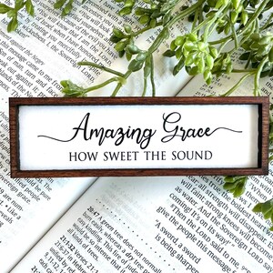 May include: A rectangular wooden sign with the words "Amazing Grace" and "How Sweet the Sound" in black script. The sign has a white background and is set against a backdrop of a Bible and greenery.