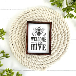 May include: A wooden sign with a brown frame and a white background. The sign has a black and white illustration of a bee and the text "Welcome to our Hive". The sign is sitting on a round, white rope coaster.