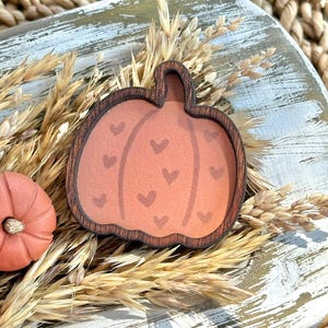 May include: A wooden pumpkin-shaped decoration with a brown frame and a peach-colored center adorned with small heart shapes. The pumpkin is surrounded by dried wheat and sits on a distressed white and gray wooden surface. Two small, orange pumpkins are also present.