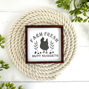 May include: A white wooden sign with a brown frame, featuring a black silhouette of a chicken and the text "FARM FRESH BUTT NUGGETS". The sign is displayed on a white surface with a beige rope circle behind it.