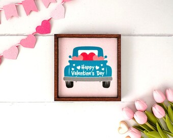May include: A square wooden-framed sign featuring a light pink background with a teal pickup truck illustration. The truck bed is filled with red and pink hearts, and the text reads Happy Valentine's Day in white lettering.
