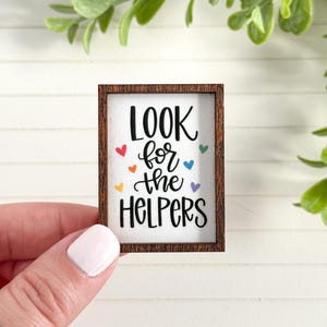 May include: A small wooden framed sign with the words "LOOK for the HELPERS" in black script. Colorful hearts in red, orange, yellow, green, blue, and purple are scattered around the text. The frame is a dark brown color.