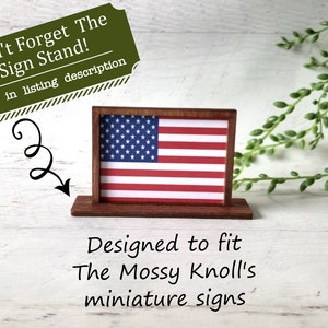 May include: A miniature wooden sign stand displaying an American flag. The sign is red, white, and blue. The text on the image says, "Don't Forget The Sign Stand!" and "Designed to fit The Mossy Knoll's miniature signs."