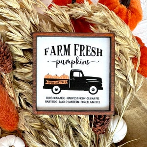 May include: A square wooden sign with the text "FARM FRESH pumpkins" and a black truck carrying orange pumpkins. The sign also lists pumpkin varieties. The sign is surrounded by dried wheat, orange and white decorative elements.