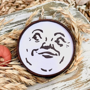May include: A round wooden ornament featuring a smiling moon face on a white background. The ornament has a dark brown wooden frame. The image is surrounded by wheat stalks and a small orange pumpkin.