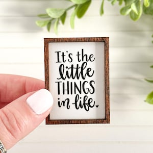 May include: Small rectangular wooden sign with a white background and a brown frame. The sign reads "It's the little THINGS in life." in black script. The sign is held by a person.