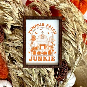 May include: A framed sign with a white background and orange text that reads "Pumpkin Patch Junkie". The sign features a stylized illustration of a barn with a windmill and pumpkins.