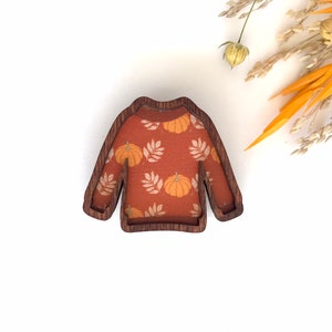 May include: A wooden cutout of a sweater with a pumpkin and leaf pattern. The sweater is brown with a light orange and white pattern.