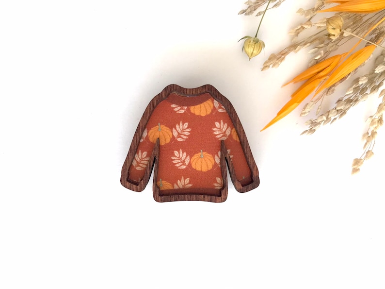 May include: A wooden cutout of a sweater with a pumpkin and leaf pattern. The sweater is brown with a light orange and white pattern.