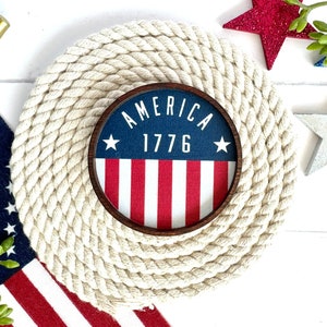 May include: A round, white rope coaster with a wooden frame and a blue, red, and white American flag design. The flag has the words "America 1776" printed on it.