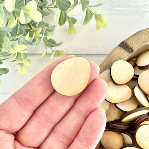 May include: A close-up of a hand holding a light brown wooden egg shape. The egg is smooth and has a slightly rounded edge. The egg is surrounded by a bowl of similar wooden shapes.